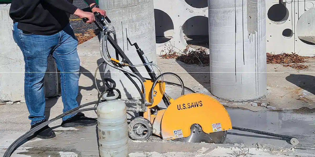 A person operating a yellow U.S. Saws walk behind saw to cut through a concrete surface, with cylindrical concrete structures in the background.
