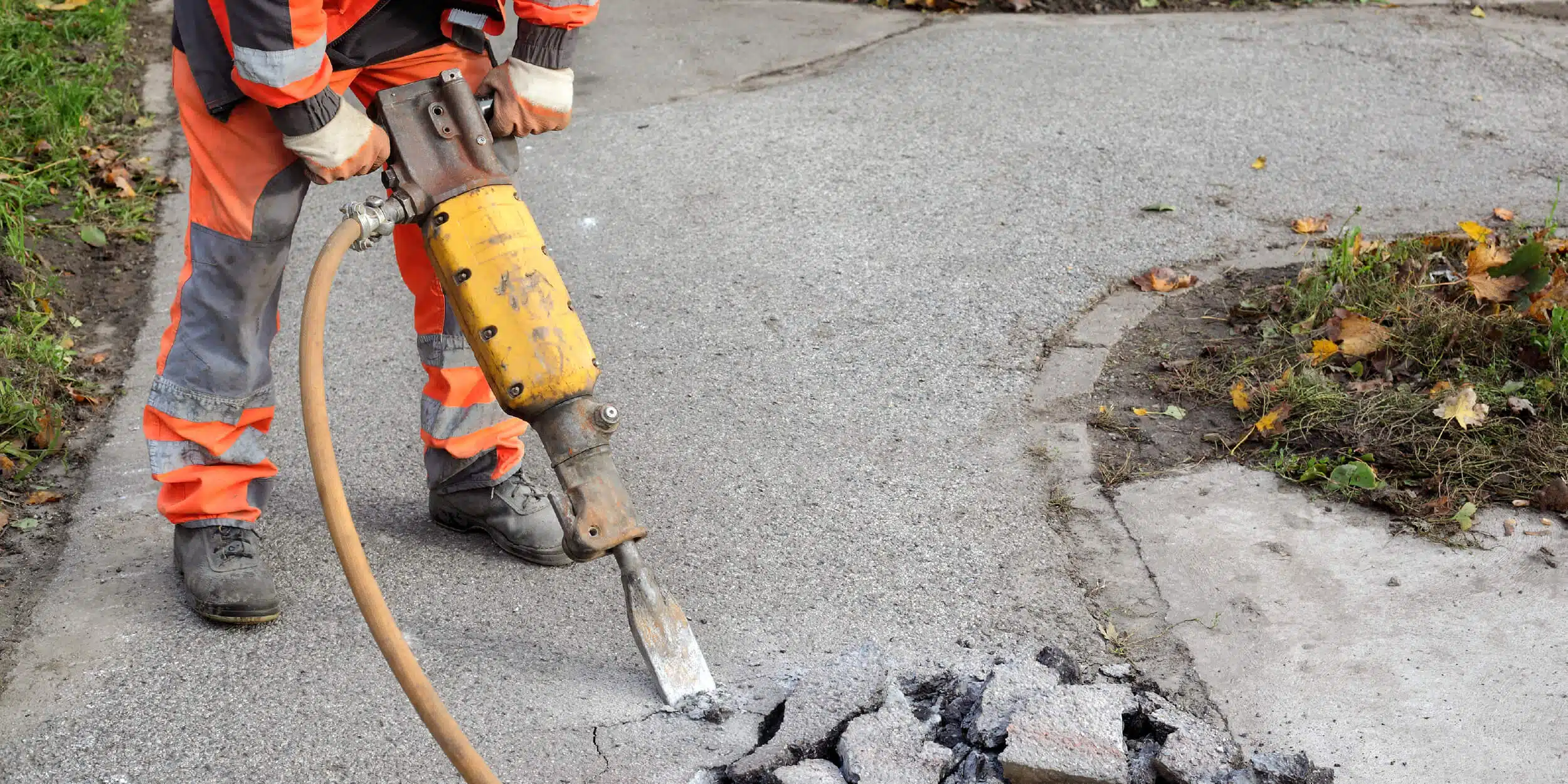 A worker in orange safety gear uses a pneumatic jackhammer to break up pavement on a sidewalk representing air saws.