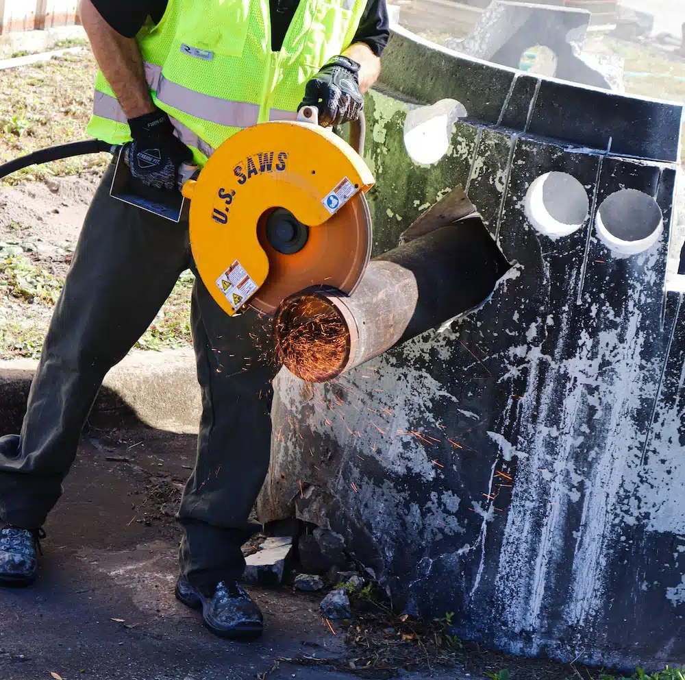A worker in a safety vest uses a power saw, similar to heavy-duty tile saws, to cut through a metal pipe, producing sparks, beside a concrete structure representing tile saws