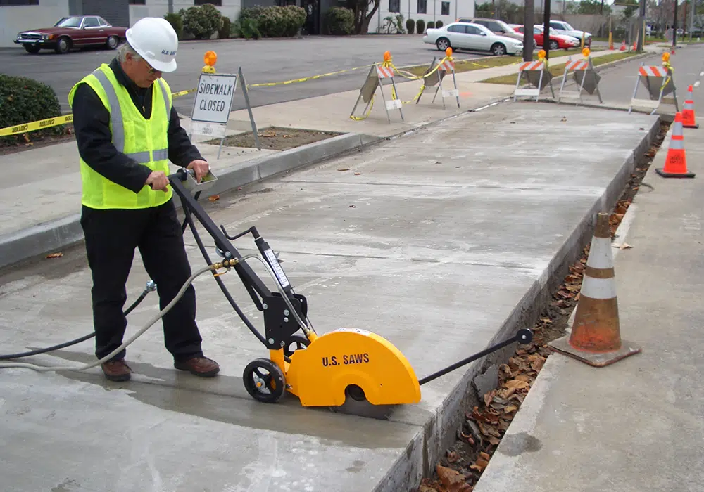 A construction worker in safety gear operates a concrete saw fitted with Turbo Rim Diamond Blades on a blocked-off sidewalk, with traffic cones and caution signs visible.