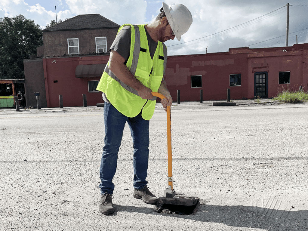 A construction worker in a safety vest and helmet performs valve maintenance while using a tool to repair a pothole on a paved surface near a red building.