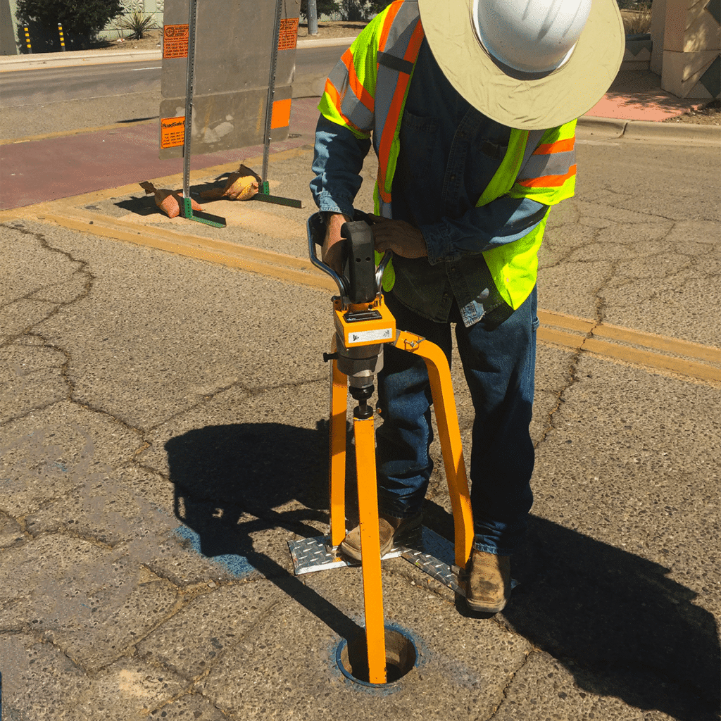 A construction worker in safety gear uses a core drill to make a circular hole in the asphalt of a road during daytime for valve maintenance.