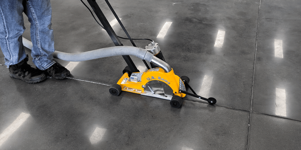 Person using a yellow concrete saw with vacuum attachment to cut a straight line on a polished garage floor for epoxy application.
